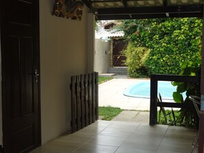 A covered patio with a pool, a door, and a view of a garden and a building in the background.