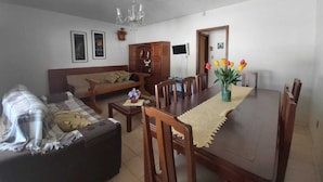 Una sala de estar con sofá, mesa de centro, mesa de comedor y sillas. Hay una araña, un televisor y una puerta con un marco de fotos.