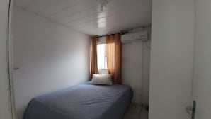 A small, single-bed room with a plain white ceiling, a window with brown curtains, and an air conditioning unit mounted on the wall. - Casa Pé na Areia, 2D, C Ar - Ingleses 41 (Florianópolis)