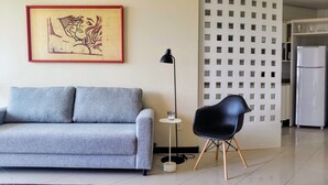 A living room with a grey sofa, a black chair, a dining table with a bowl, and a kitchen area with a fridge. - Ap Premium na Lagoa (Florianópolis)