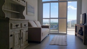 A living room with a sofa, a television on a wooden stand, and a view through large windows. - Cobertura Liberty Campeche (Florianópolis)