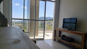 A room with a large window, a wooden bench, a TV on a wooden stand, and a view of a balcony with mountains in the background. - Cobertura Liberty Campeche (Florianópolis)