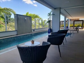 Uma área à beira da piscina com vista para a piscina, cadeiras de vime, uma mesa pequena e um vaso de plantas.