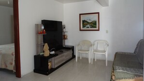 Ein Wohnzimmer mit Fernseher, zwei weißen Sessel, einer Couch und einem Gemälde an der Wand.