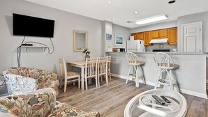 A modern living room with a flat-screen TV, a dining area with wooden chairs, a kitchen with wooden cabinets, and a round glass table with a remote control.