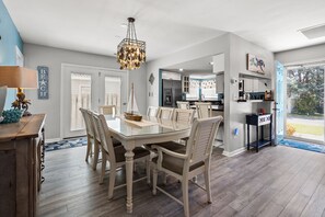 Un comedor amplio con una mesa de vidrio y sillas blancas, un candelabro, y vistas a la cocina y al exterior.