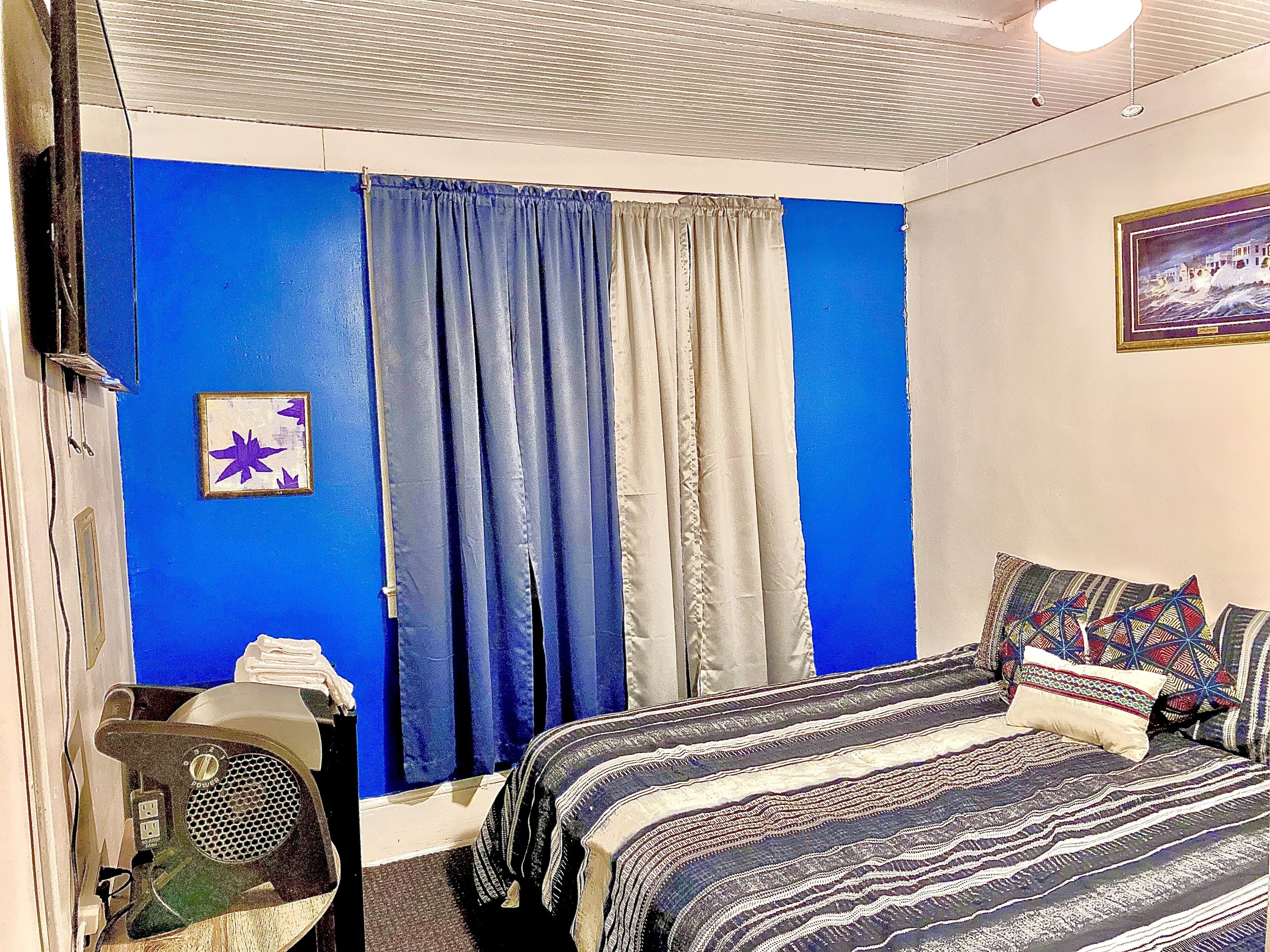 Un dormitorio con una decoración azul y blanca, una cama con ropa de cama estampada, una mesita con un ventilador y un cuadro en la pared.