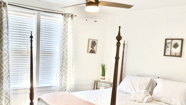 A neatly made bed with a wooden frame, a ceiling fan, and a window with blinds. - Bella's Boutique Hotel (Hemingway)