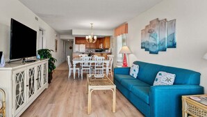 Un salon avec un canapé bleu, une table à manger avec des chaises, un téléviseur posé sur un meuble, et une cuisine en arrière-plan.