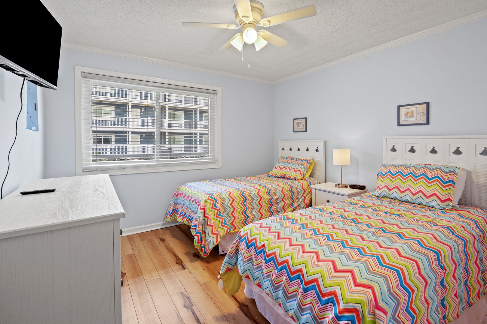 A bedroom with two beds, a desk, a ceiling fan, and a window with blinds.