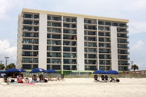 Un hôtel à plusieurs étages doté de balcons, situé sur une plage de sable, avec des chaises longues et des parasols.