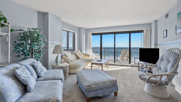 A spacious living room with a large window overlooking the ocean, a comfortable sofa, a coffee table, and a wicker chair. - Panoramic ocean views, pool, hot tub, close to shops, restaurants and Cherry Grove Beach (North Myrtle Beach)
