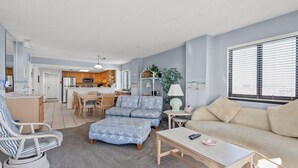 A modern living room with a sofa, armchair, and ottoman, a dining area with a table and chairs, and a kitchen with wooden cabinets and appliances. - Panoramic ocean views, pool, hot tub, close to shops, restaurants and Cherry Grove Beach (North Myrtle Beach)