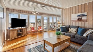 A spacious living room with a large TV, wooden furniture, a sofa, and a view through large windows. - Oceanfront, dog-friendly vacation home w/gorgeous ocean views, large deck-steps from beach (North Myrtle Beach)