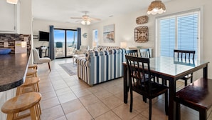 A spacious living area with a striped sofa, a dining table with chairs, a kitchen area with a counter and stools, and a ceiling fan.