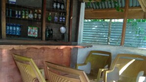 Una zona de bar con sillas de plástico y una mesa de madera, una estantería con botellas y una ventana con persianas.