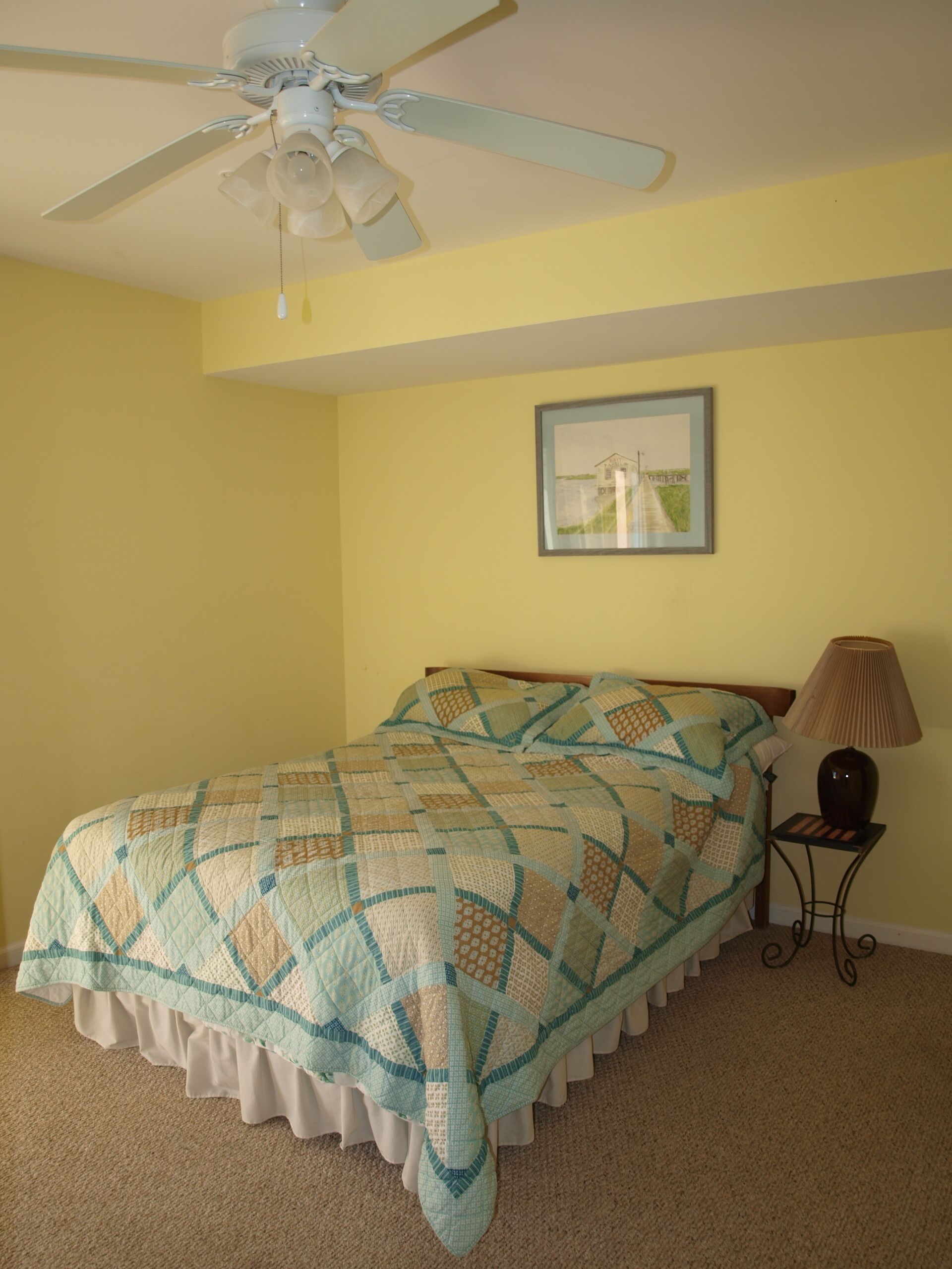 A bedroom with a bed, a ceiling fan, a framed picture on the wall, and a bedside lamp.