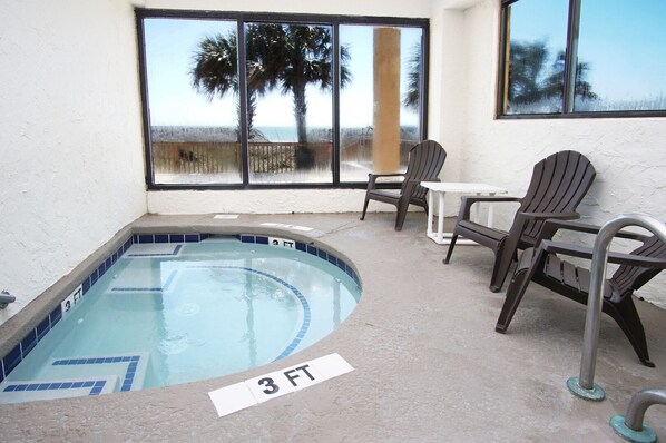 A small indoor pool with a 3 FT depth marker, surrounded by patio chairs and a table, with a view of palm trees and the beach through large windows.