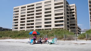 A multi-story building with numerous balconies, situated near a sandy area with beach chairs and an umbrella.