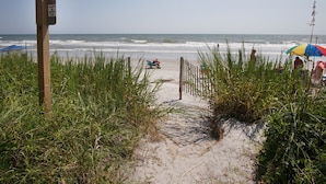 A sandy beach path leading to the ocean, with a wooden signpost and a beach umbrella in the background.