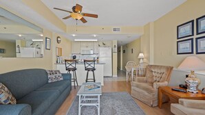 A living room with a sofa, a coffee table, and a chair. There is a kitchen in the background with a refrigerator, microwave, and chairs. - Rest, pure & simple! Gorgeous ocean view, large balcony w/pool & hot tub in popular Cherry Grove (North Myrtle Beach)