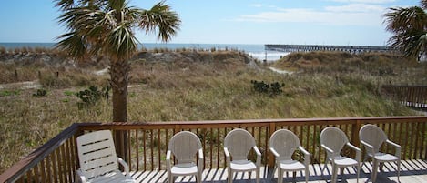 Une terrasse en bois avec des chaises blanches et un banc, donnant sur une plage et une jetée.