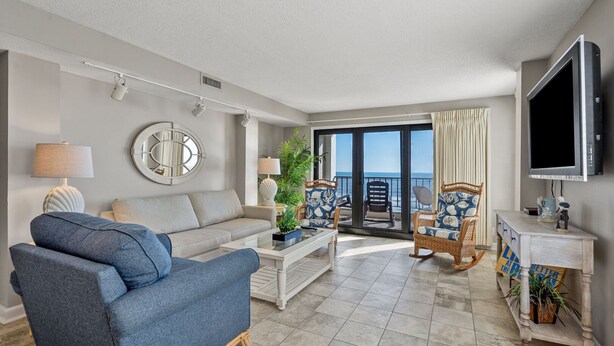 A living room with a sofa, coffee table, and chairs, a television, and a view of the ocean through large glass doors. - This view! You'll love this roomy oceanfront condo rental w/pool, sundeck in popular North Myrtle (North Myrtle Beach)