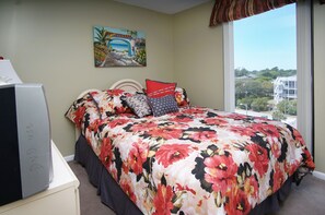 A bedroom with a floral bedspread, a painting of a coastal scene, and a view of buildings through a large window.