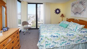 Habitación de hotel con una cama grande, un aparador de madera, una silla, un ventanal con vistas a edificios y un reloj de pared.