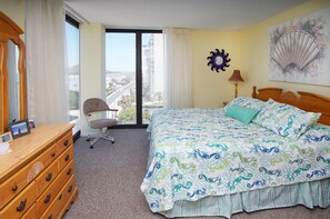 A hotel room with a large bed, a wooden dresser, a chair, a window with a view of buildings, and a wall clock.
