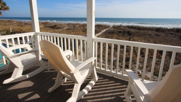 Een houten terras met witte leuningen en twee schommelstoelen, gericht op het strand.