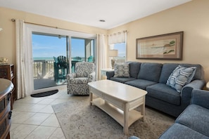 A living room with a sofa, armchair, and coffee table, a balcony with chairs, and a painting on the wall.