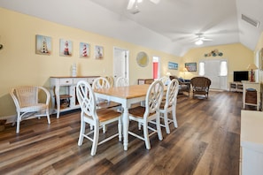 Uma sala de jantar com mesa de madeira e cadeiras brancas, uma cozinha com armários brancos e uma sala de estar com móveis de vime.