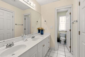 Une salle de bain avec deux lavabos, un grand miroir et des toilettes visibles à travers une porte ouverte.