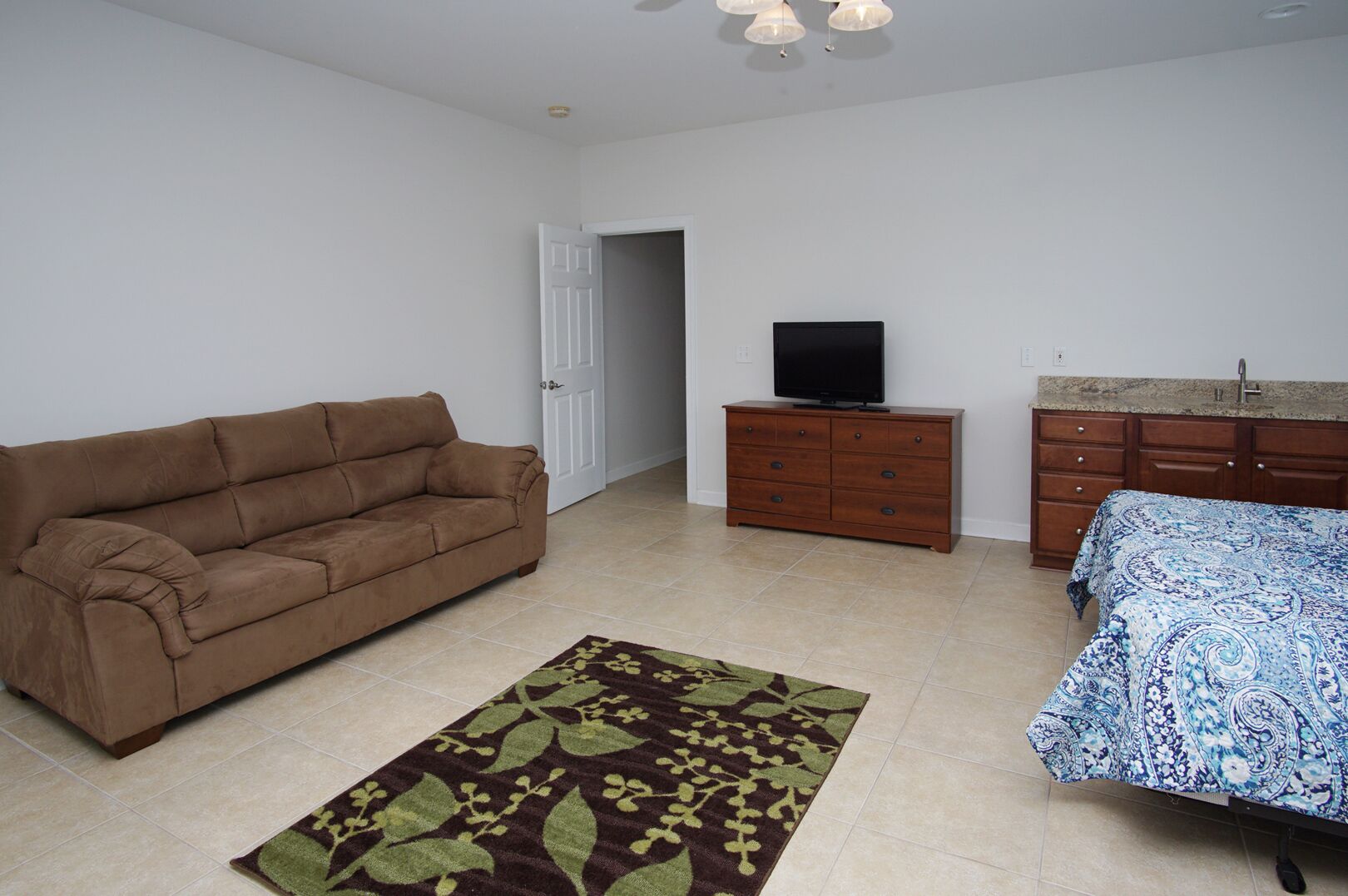 Una sala de estar con un sofá marrón, un televisor sobre un mueble de madera, un tapete estampado y una cama con colcha azul y blanca.