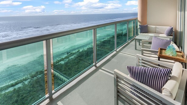 A balcony with a glass railing overlooking the ocean, featuring a sofa and chairs with striped cushions. - Brilliant oceanfront condo w/indoor & outdoor pools, lazy river, fitness center, kids' pool (North Myrtle Beach)