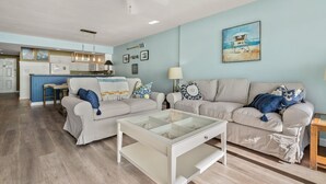 A spacious living area with a light blue wall, a beige sofa, a glass coffee table, and a kitchen in the background. - Brilliant oceanfront condo w/indoor & outdoor pools, lazy river, fitness center, kids' pool (North Myrtle Beach)