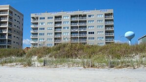 A multi-story residential building with balconies, located near a sandy beach with a fence and vegetation. - Oceanfront panoramic views, indoor pool, Jacuzzi, balcony--in favorite North Myrtle Beach (North Myrtle Beach)