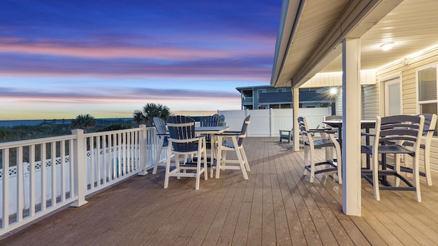 Una terraza de madera con mobiliario exterior y barandilla, con vista a una playa.