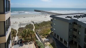 A beach view with a pier extending into the ocean, a sandy beach, and a building with a flat roof and multiple air conditioning units. - Charming & comfortable condo with ocean view, balcony, w/pool & hot tub in popular Cherry Grove (North Myrtle Beach)