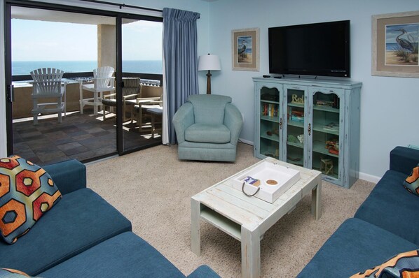 A living room with a sofa, armchair, and a coffee table. There is a TV mounted on the wall and a bookshelf. - Charming & comfortable condo with ocean view, balcony, w/pool & hot tub in popular Cherry Grove (North Myrtle Beach)