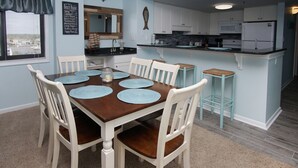 A dining area with a wooden table and white chairs, a kitchen with white cabinets and a refrigerator, and a window with blinds. - Charming & comfortable condo with ocean view, balcony, w/pool & hot tub in popular Cherry Grove (North Myrtle Beach)