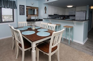 A dining area with a wooden table and white chairs, a kitchen with white cabinets and a refrigerator, and a window with blinds. - Charming & comfortable condo with ocean view, balcony, w/pool & hot tub in popular Cherry Grove (North Myrtle Beach)