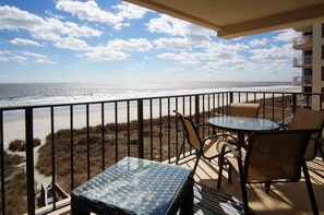 A balcony with a glass table and chairs, overlooking a beach with dunes and a clear sky. - Stylish oceanfront condo w/pool, kid's pool and  private deck for sunrises & sunsets (North Myrtle Beach)