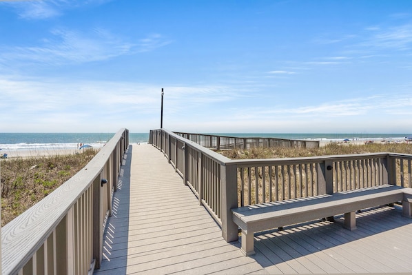 A wooden boardwalk leading to a beach with a bench and a lamp post.