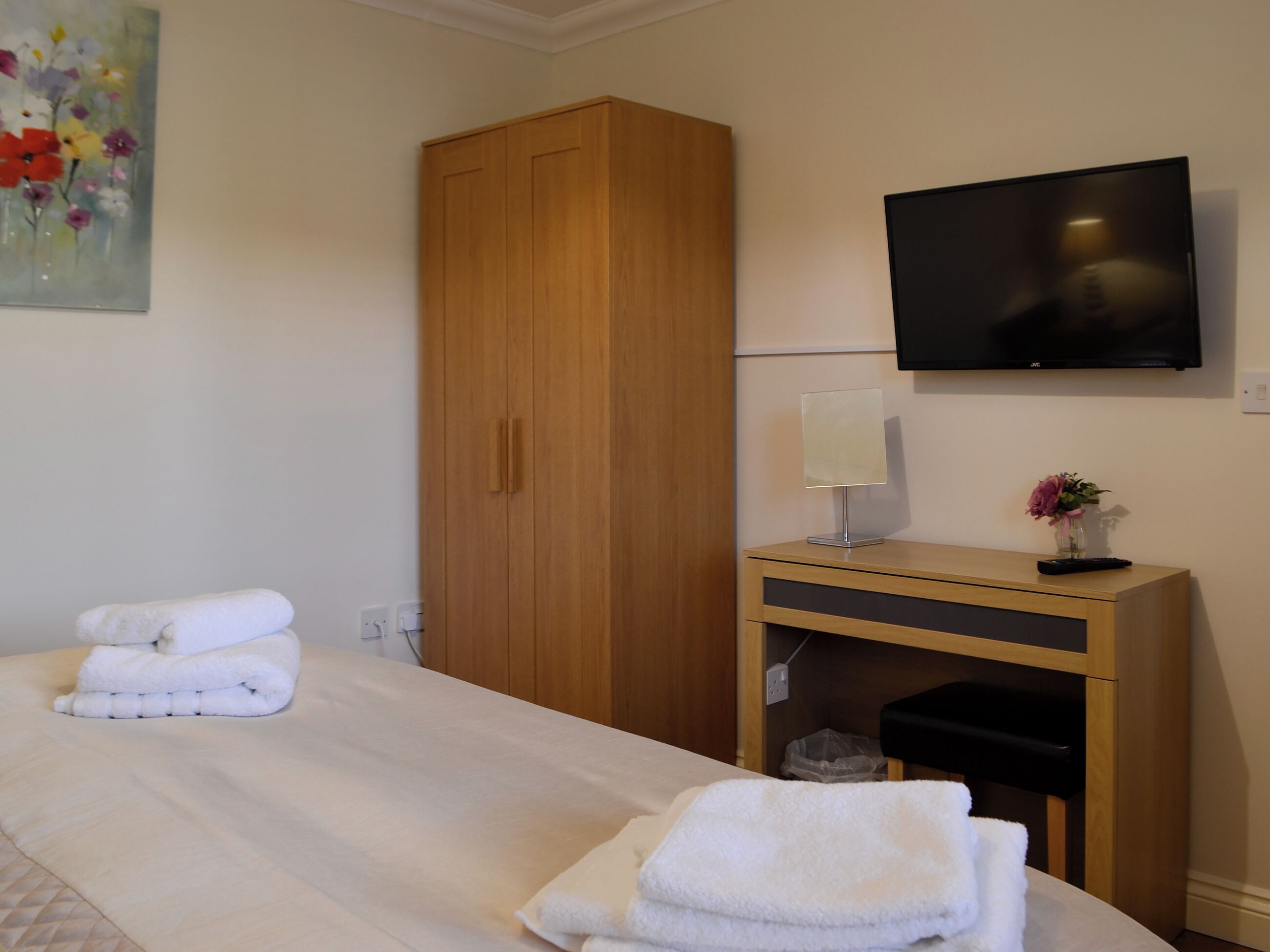 A hotel room with a bed, a wooden wardrobe, a television, a desk with a lamp, and a painting on the wall.