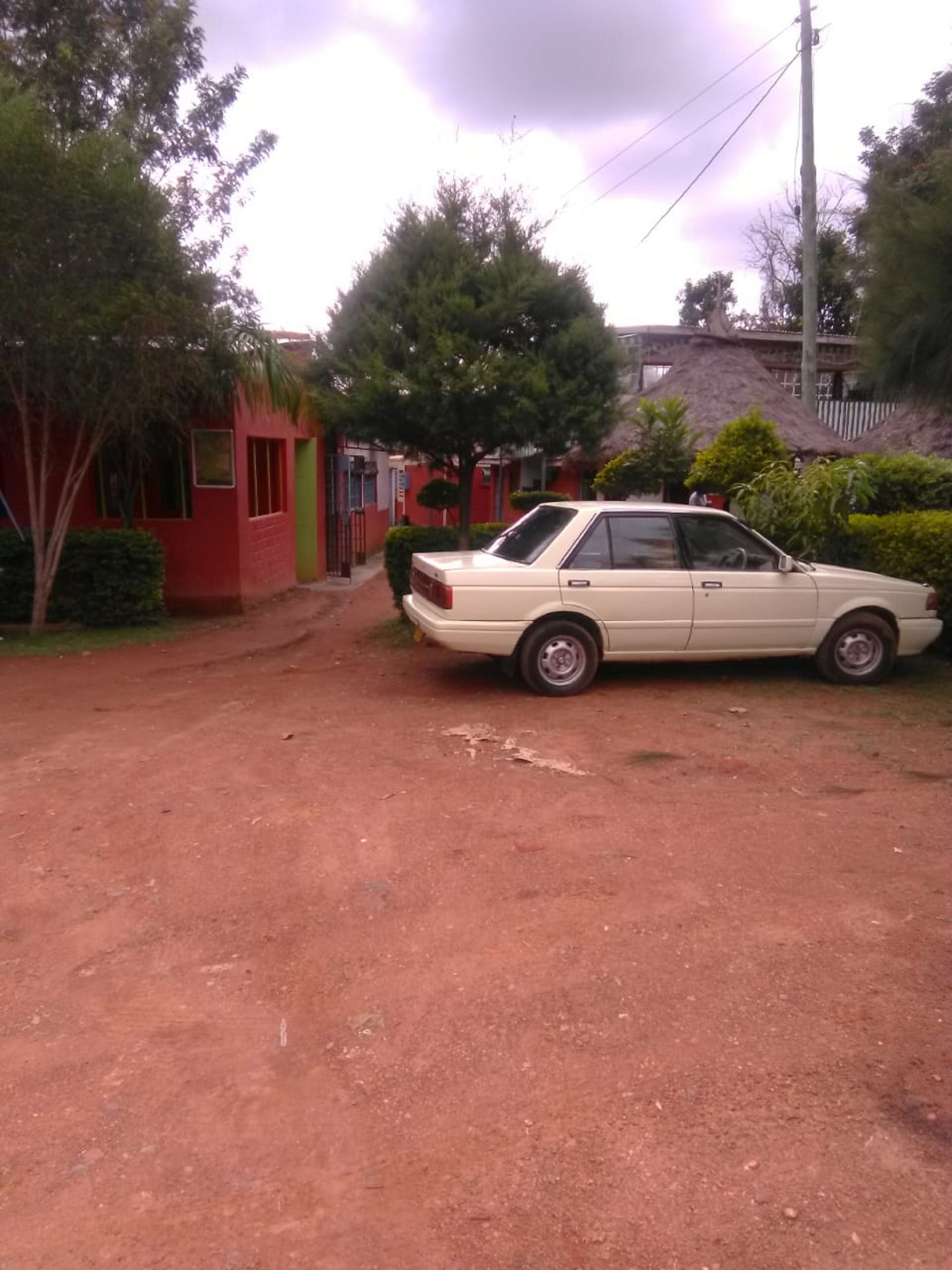 Un coche color arena estacionado en un camino de tierra en una zona residencial con edificios rojos y verdes, rodeado de árboles y una construcción con techo de paja.