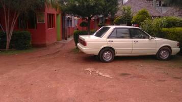 Un coche color arena estacionado en un camino de tierra en una zona residencial con edificios rojos y verdes, rodeado de árboles y una construcción con techo de paja.