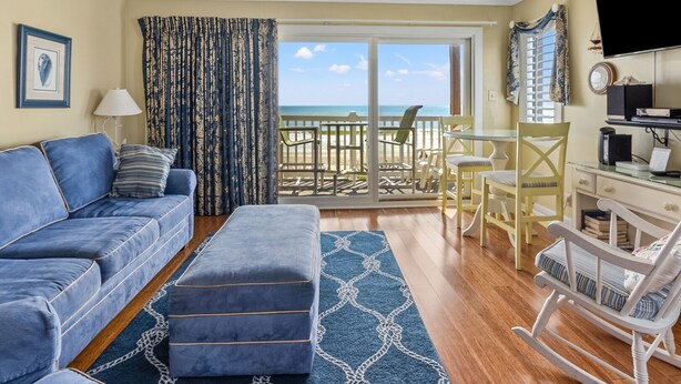 A living room with a blue sofa, ottoman, and a dining area with chairs and a table. - Charming oceanfront condos w/pool, price-friendly in perfect area - North Myrtle Beach rental (North Myrtle Beach)