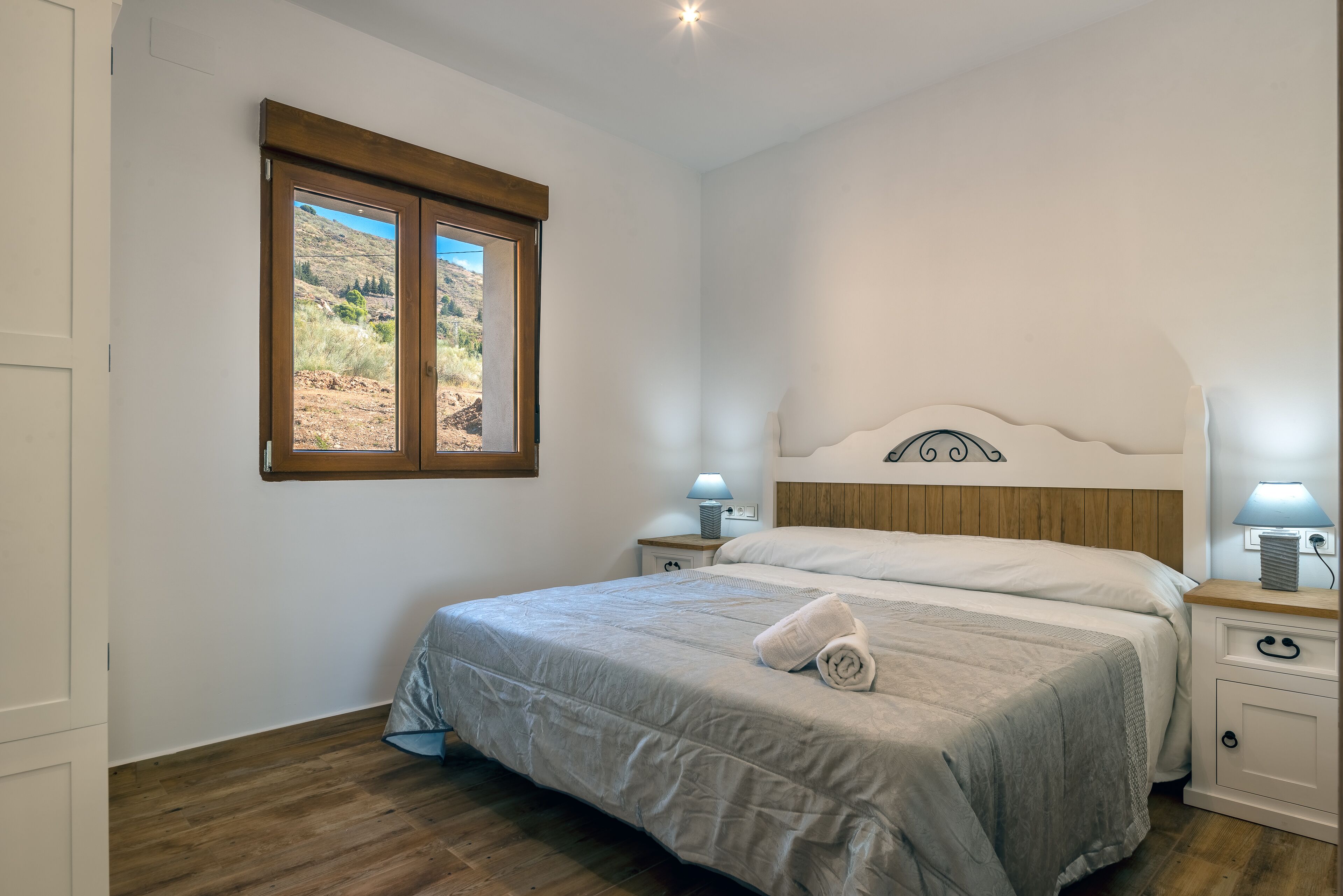 Un dormitorio con cama, dos mesitas de noche con lámparas, una ventana con vistas a un paisaje montañoso y suelo de madera.